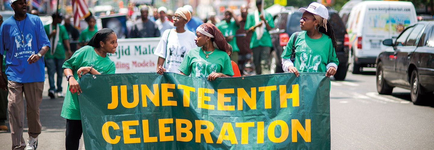 People march in a parade while holding a Juneteenth celebration banner.