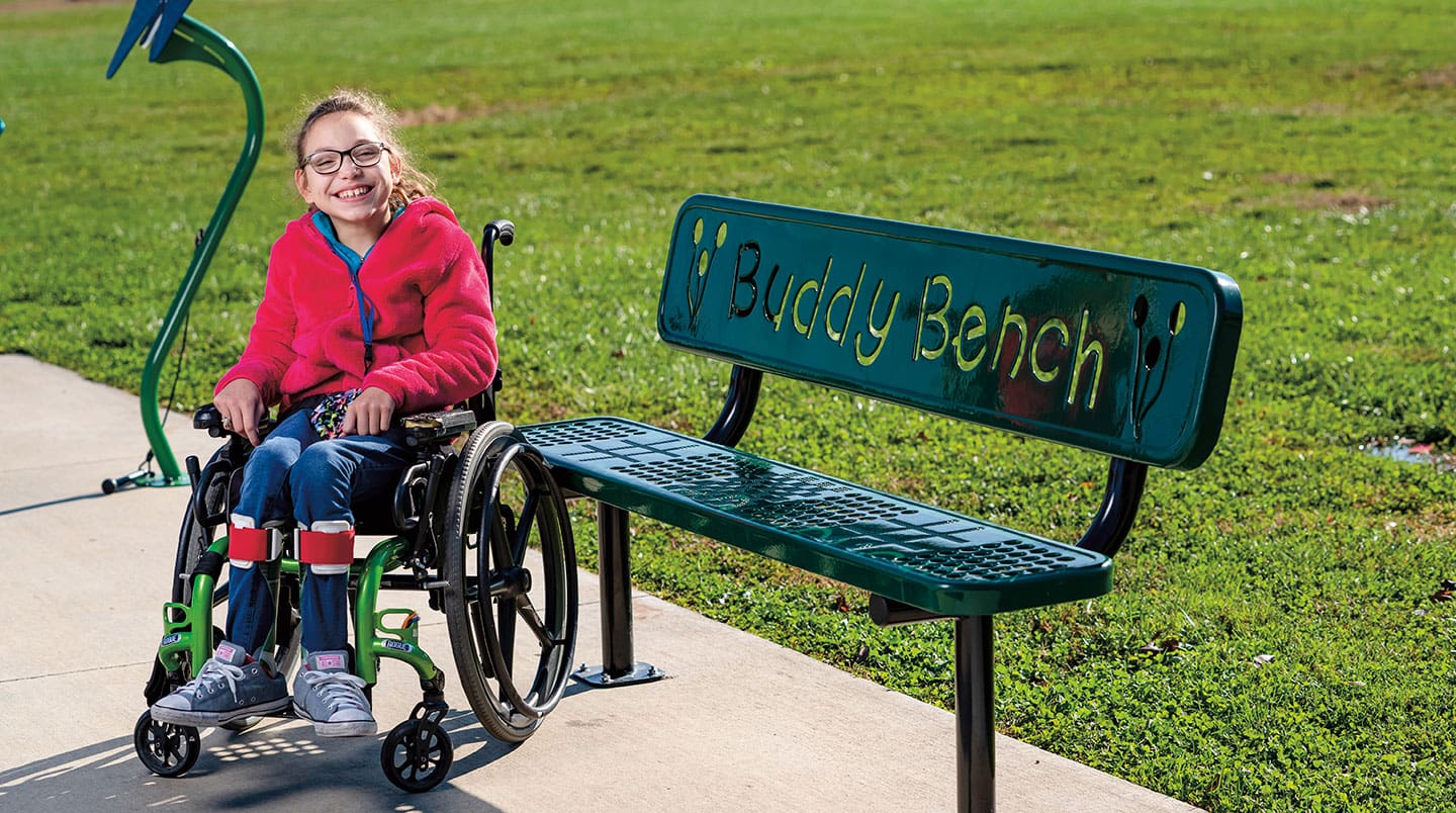 Melody Day smiles and sits in a while chair next to a buddy bench