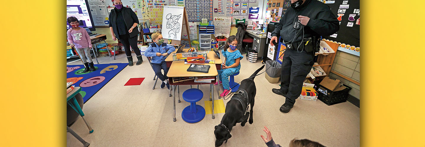 An officer holds a sniffing dog’s leash in a classroom.