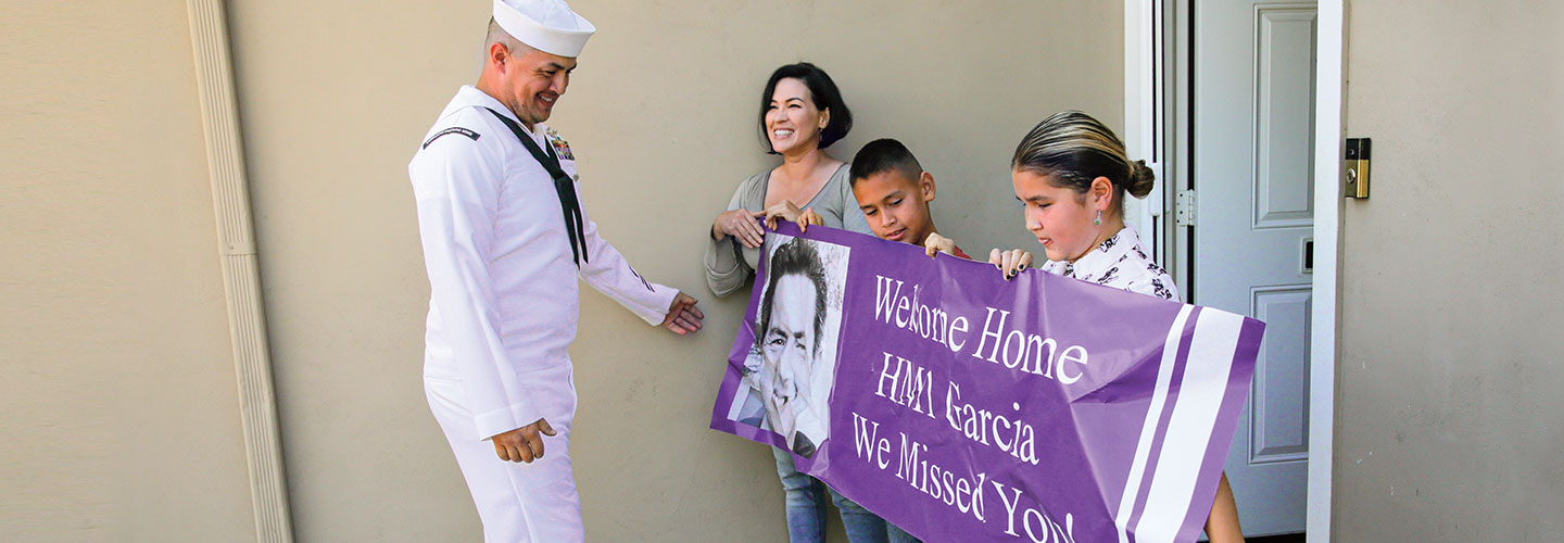 A man in a white naval sailor uniform being welcomed home by his family 