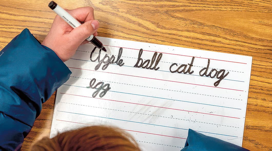A child writes cursive words apple, ball, cat, dog, and egg on lined paper at a desk