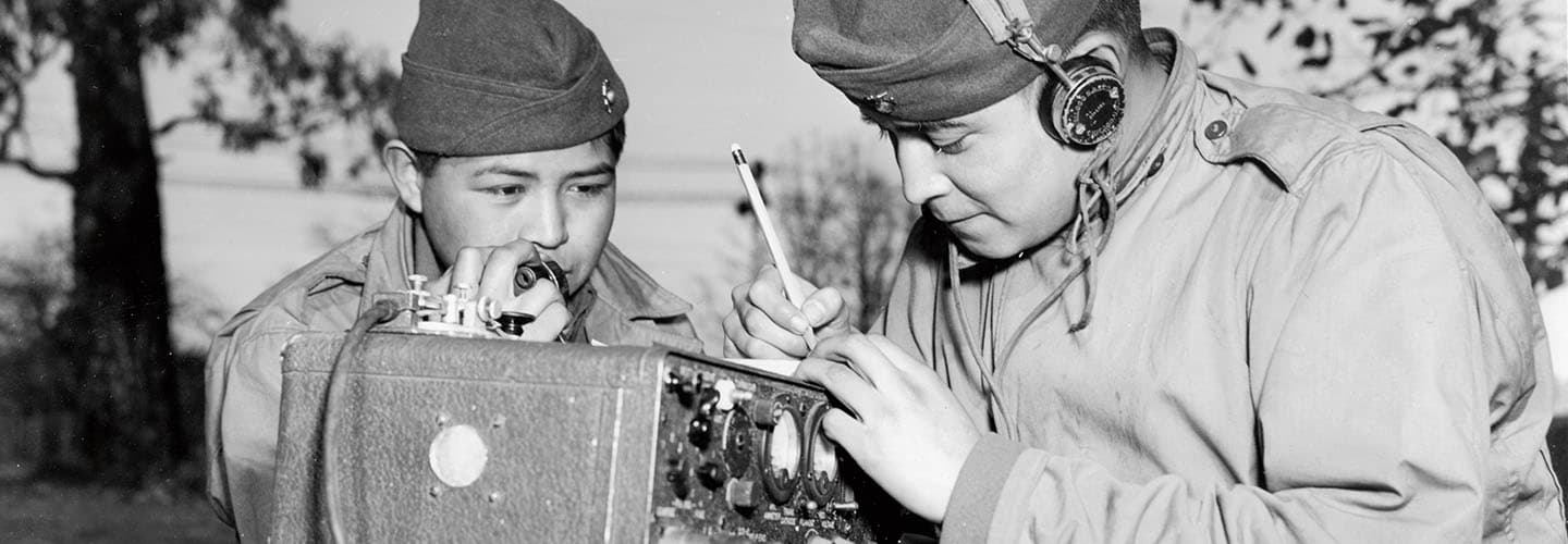 Historical photo of a soldier speaking into a field radio handset as another in headphones writes