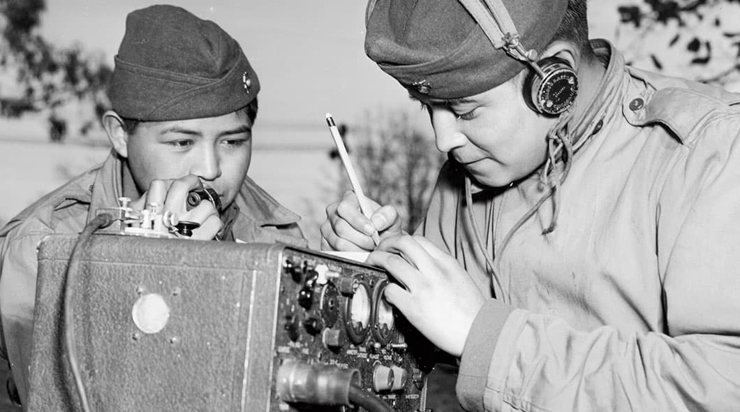 Historical photo of a soldier speaking into a field radio handset while another writes with a pencil